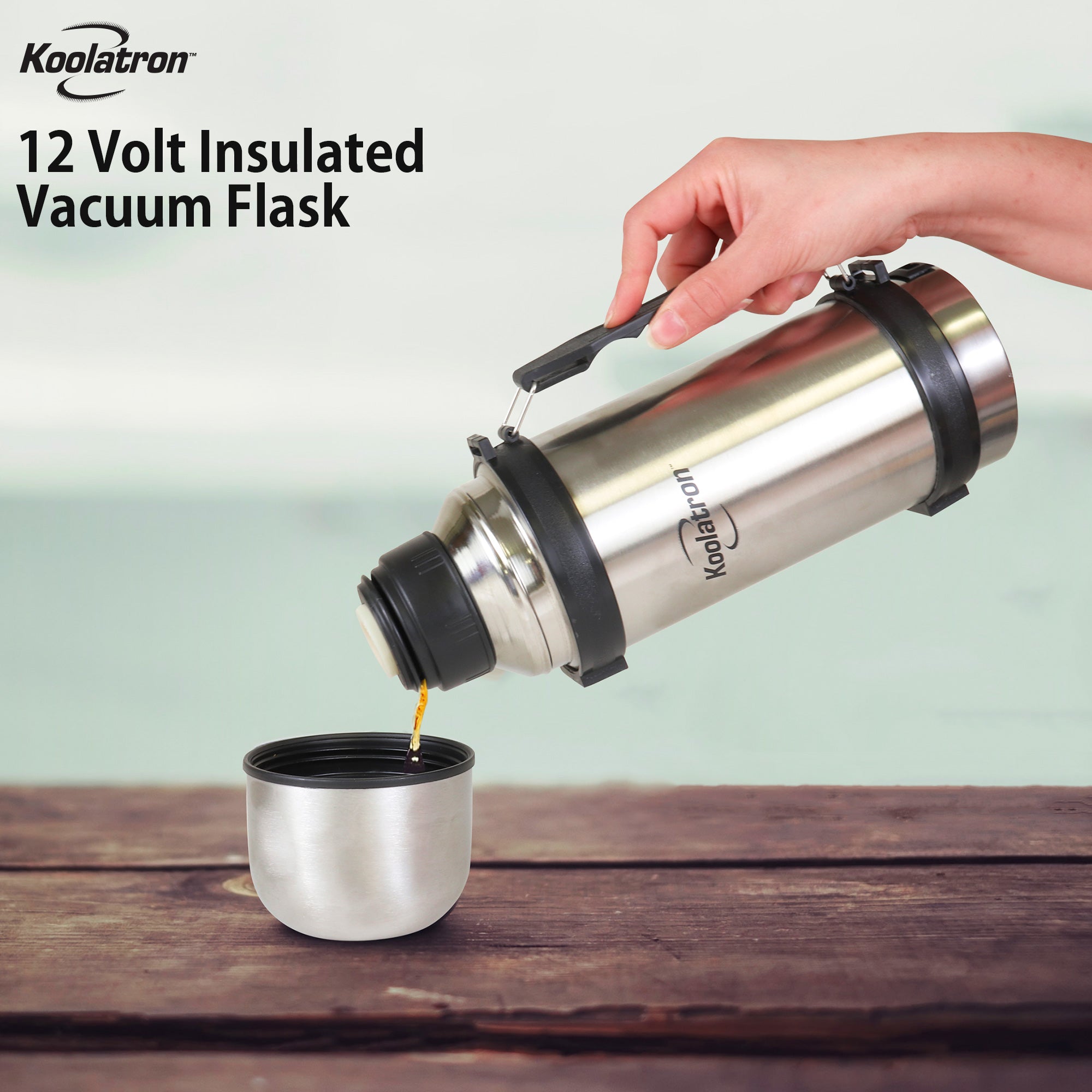 Lifestyle image of a person's hand holding the vacuum flask by its handle and pouring coffee into the single serve cup lid. The cup is sitting on a dark brown wood surface and the background is light blue. Text overlay reads Kool-atron12 Volt insulated vacuum flask.