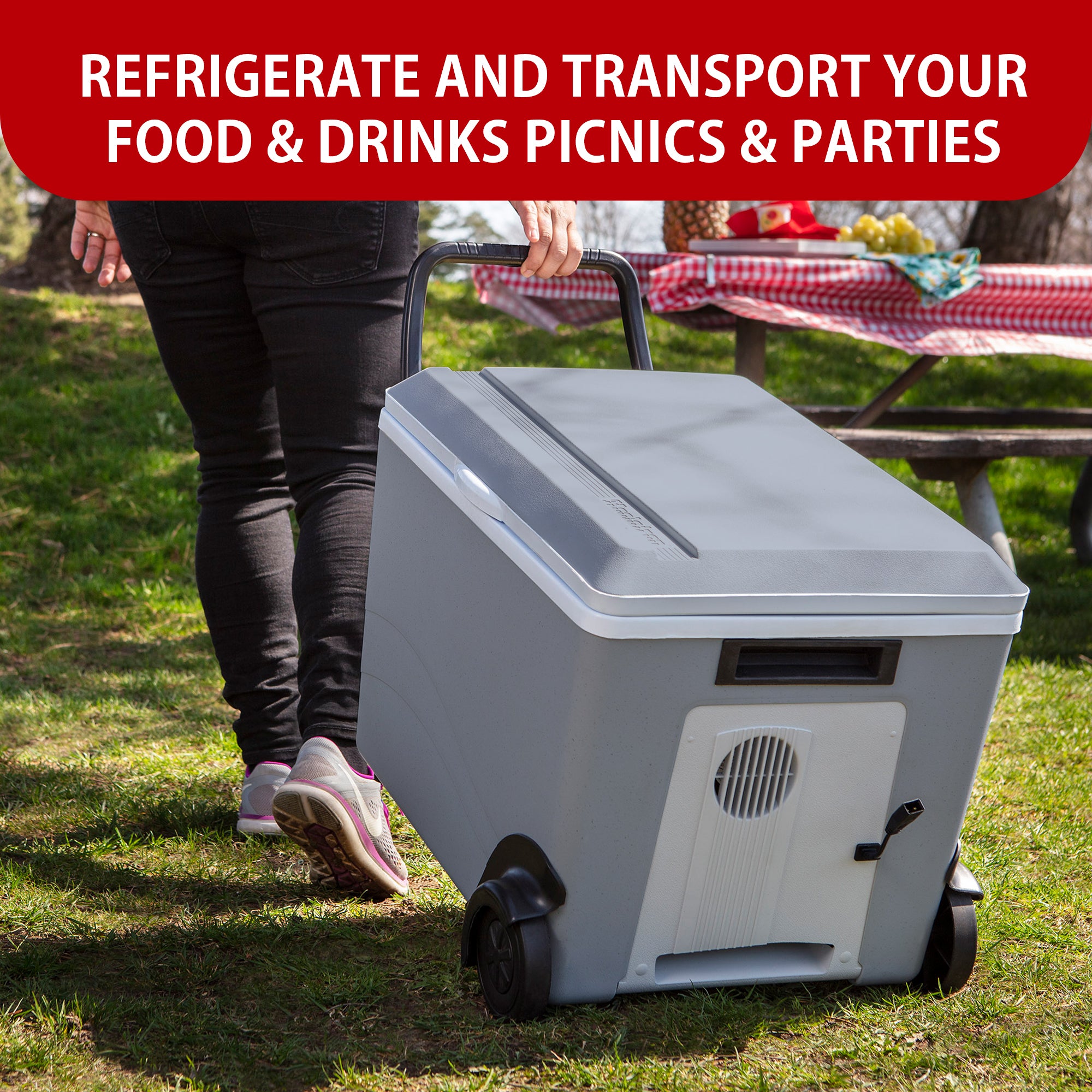 A person wearing black pants and gray and pink running shoes is towing the Kool-atronrolling 12V cooler/warmer on grass towards a picnic table with a red and white checked tablecloth. Text above reads, "REFRIGERATE AND TRANSPORT FOOD AND DRINKS TO PICNICS AND PARTIES"