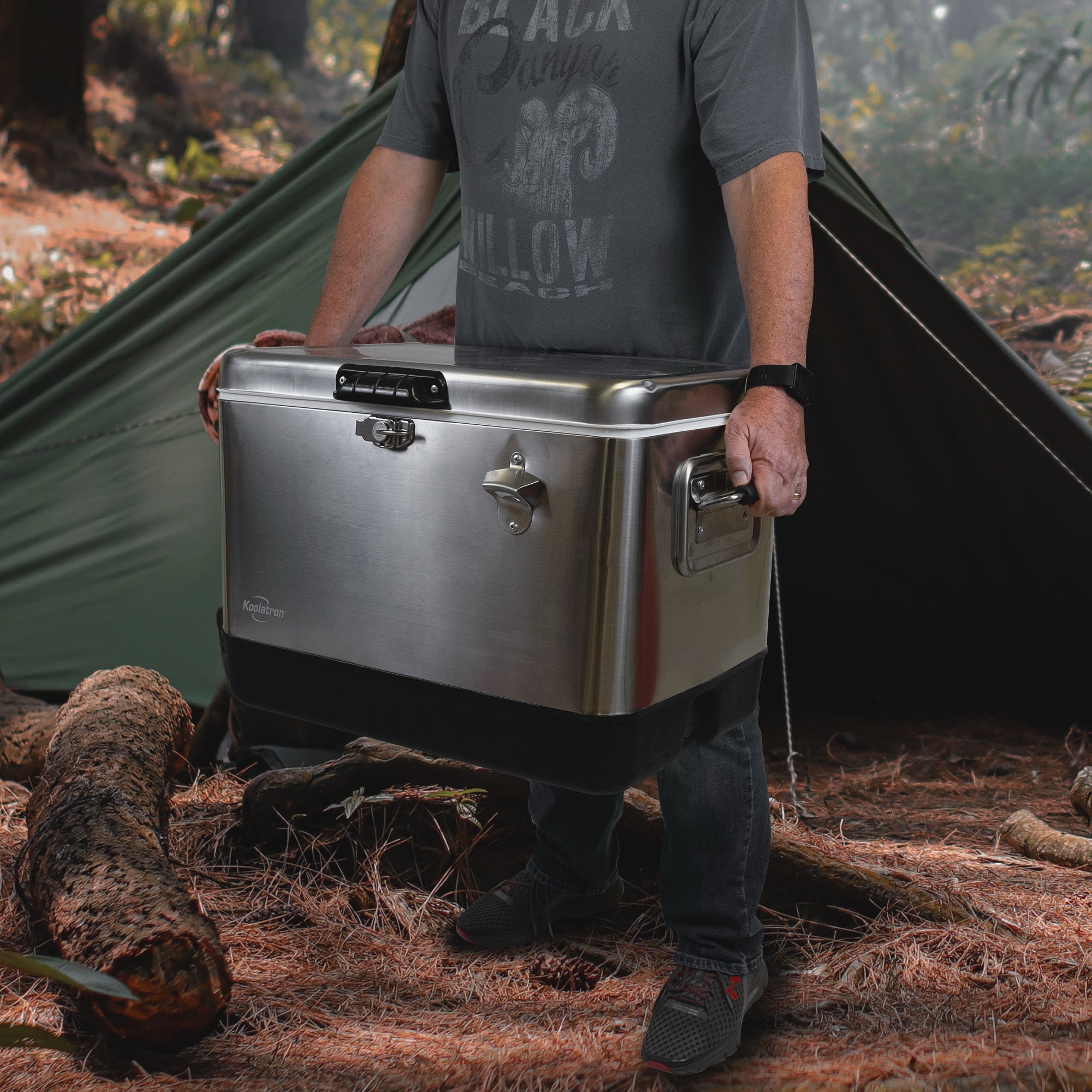 Lifestyle image of a person wearing jeans and a blue t-shirt standing up holding the Kool-atronstainless steel 54 quart ice chest in front of a dark green A-frame tent