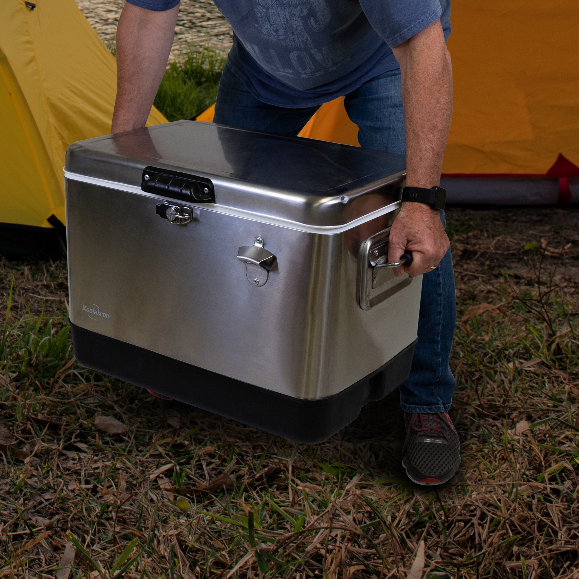 Lifestyle image of a person wearing jeans and a blue t-shirt crouching to pick up the Kool-atronstainless steel 54 qt ice chest in front of two yellow dome tents