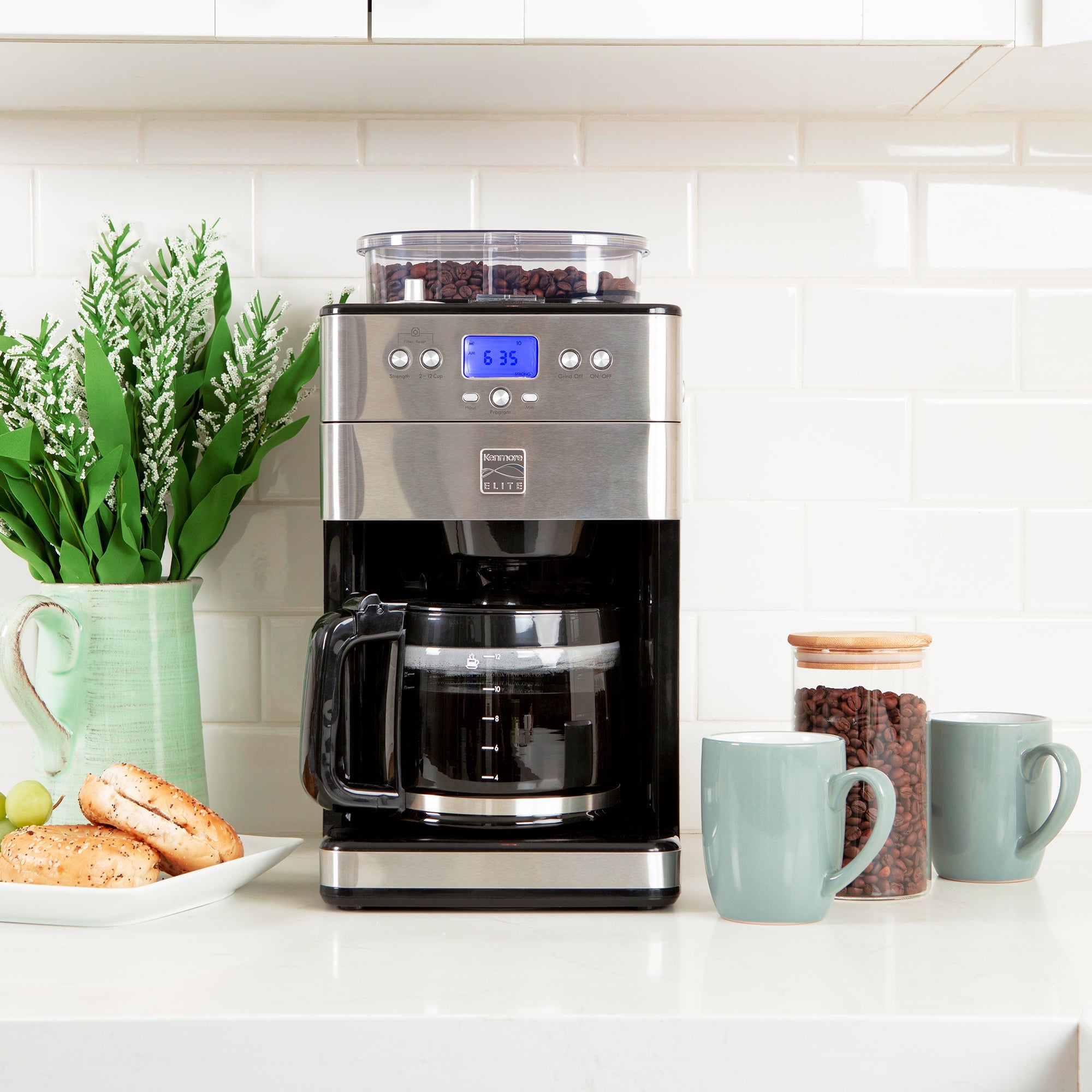 Kenmore grind and brew coffee maker filled with brewed coffee on a white quartz countertop with white tile backsplash. There is greenery in a ceramic pitcher and a plate with sliced bagels to the left and a glass jar of coffee beans and two light blue mugs to the right