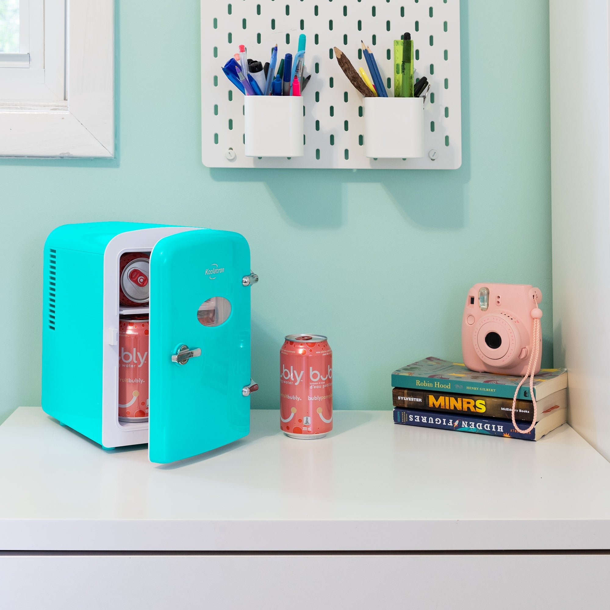 Lifestyle image of Kool-atronretro 6 can mini fridge, partly open with cans inside, on a white desktop with an aqua wall behind. There is a can of soda, a stack of books, and a pink camera to the right of the fridge and two cups of pens and pencils attached to the wall above
