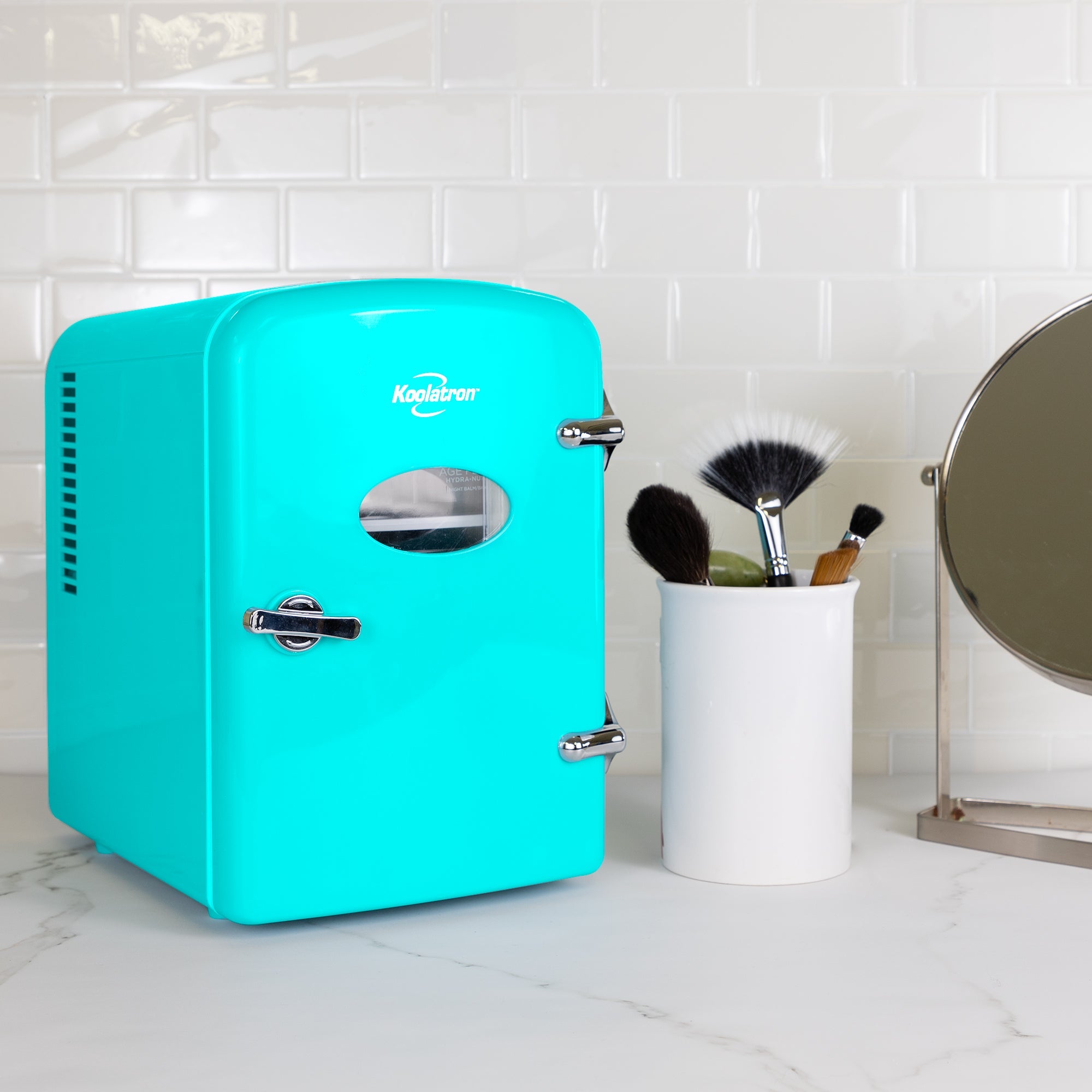 Lifestyle image of Kool-atronretro 6 can mini fridge, closed, with makeup brushes in a white container on its right, on a white marbled counter with a white tile backsplash behind 