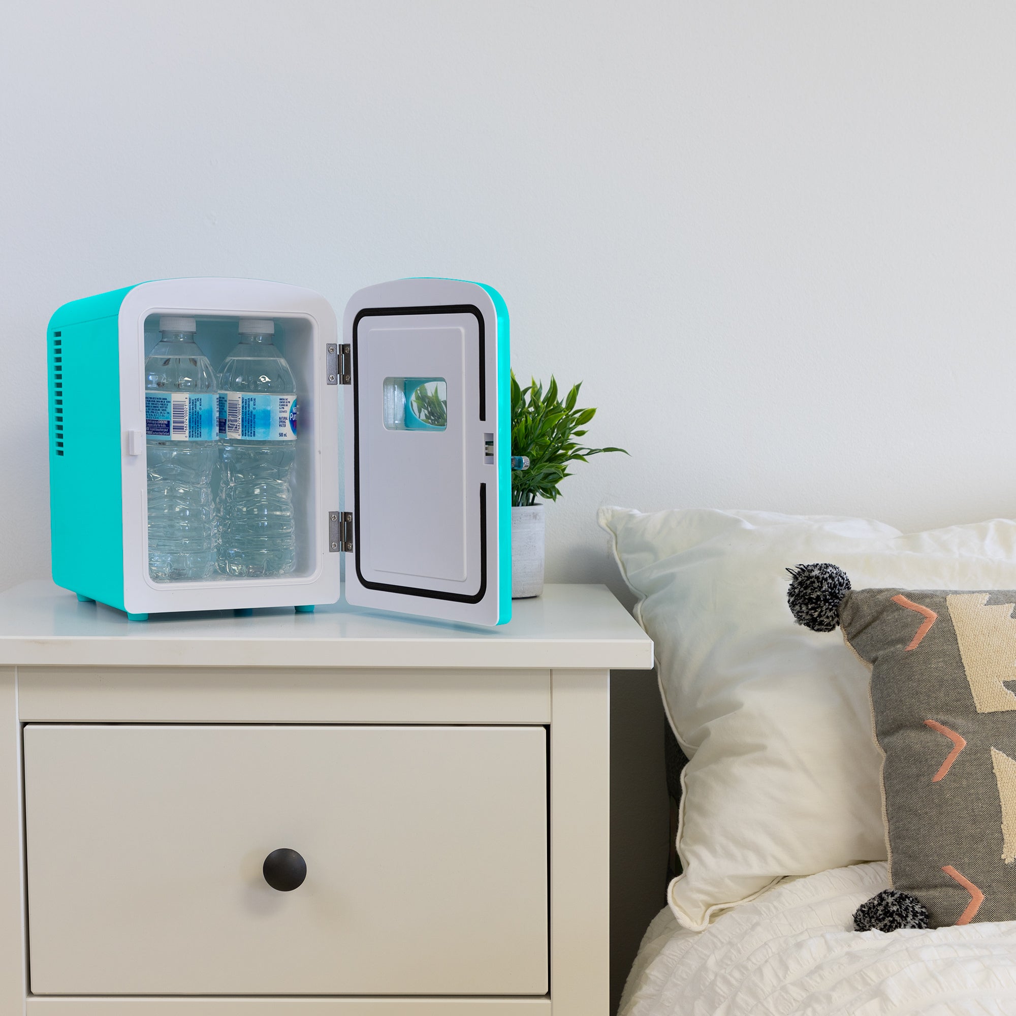 Lifestyle image of Kool-atronretro 6 can mini fridge, open with 4 water bottles inside, with a plant in a white pot on its right, on a white bedside table beside a bed with white linens