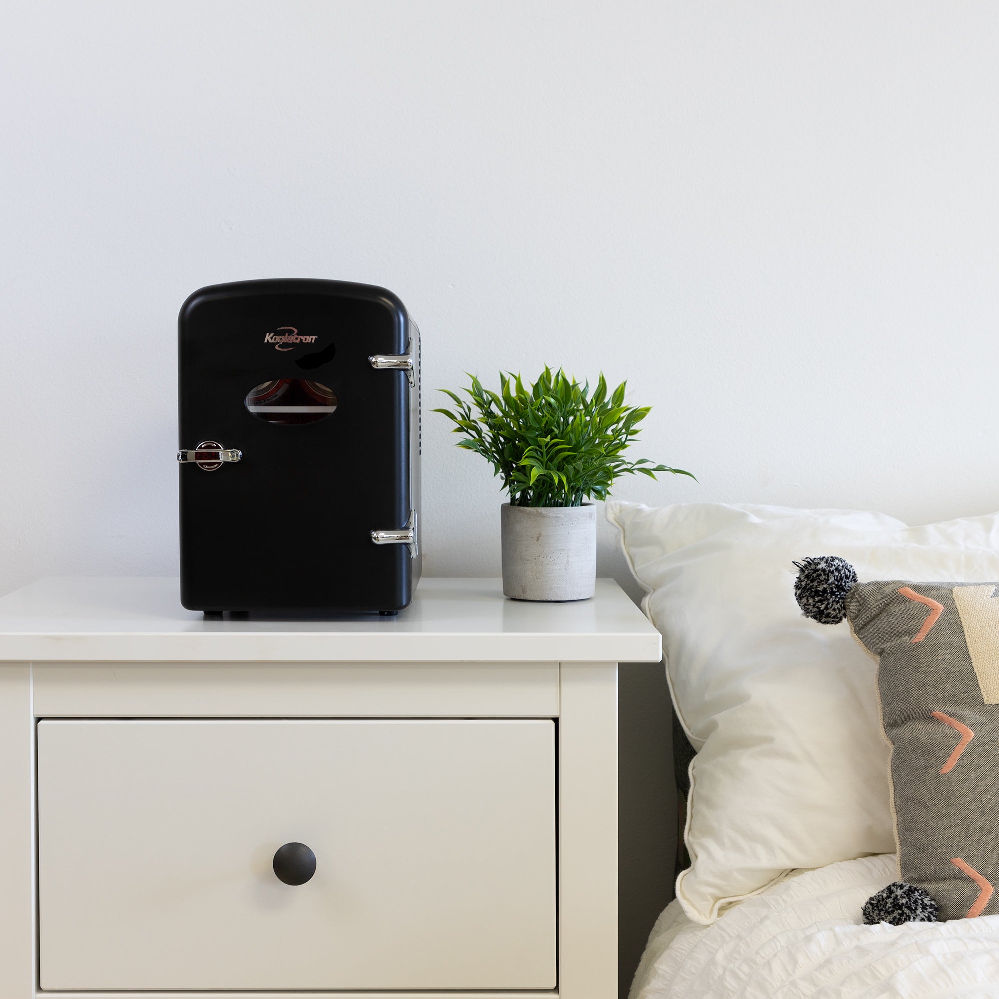 Lifestyle image of Kool-atronretro 6 can mini fridge, closed, with a plant in a white pot on its right, on a white bedside table beside a bed with white linens 