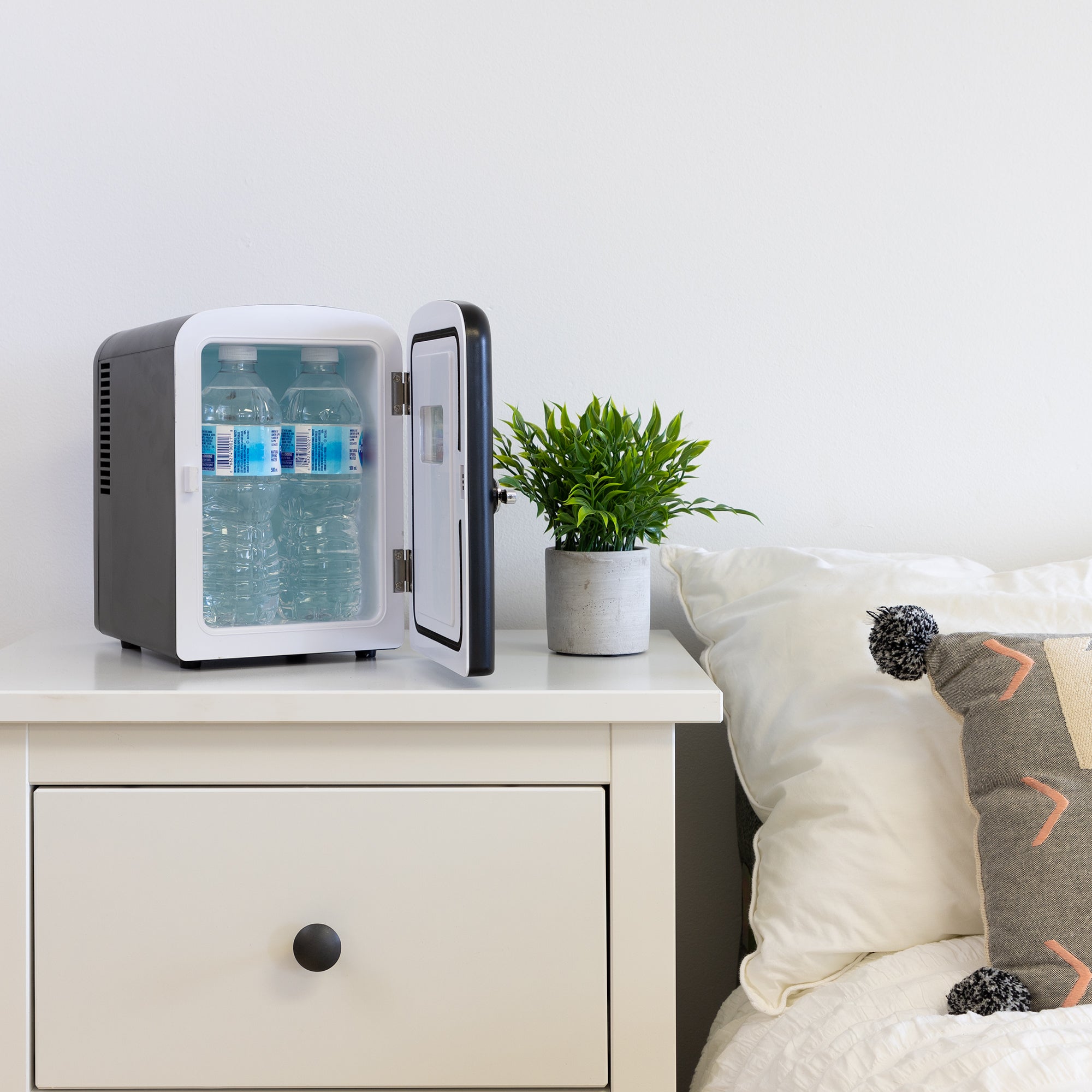 Lifestyle image of Kool-atronretro 6 can mini fridge, open with 4 water bottles inside, with a plant in a white pot on its right, on a white bedside table beside a bed with white linens 