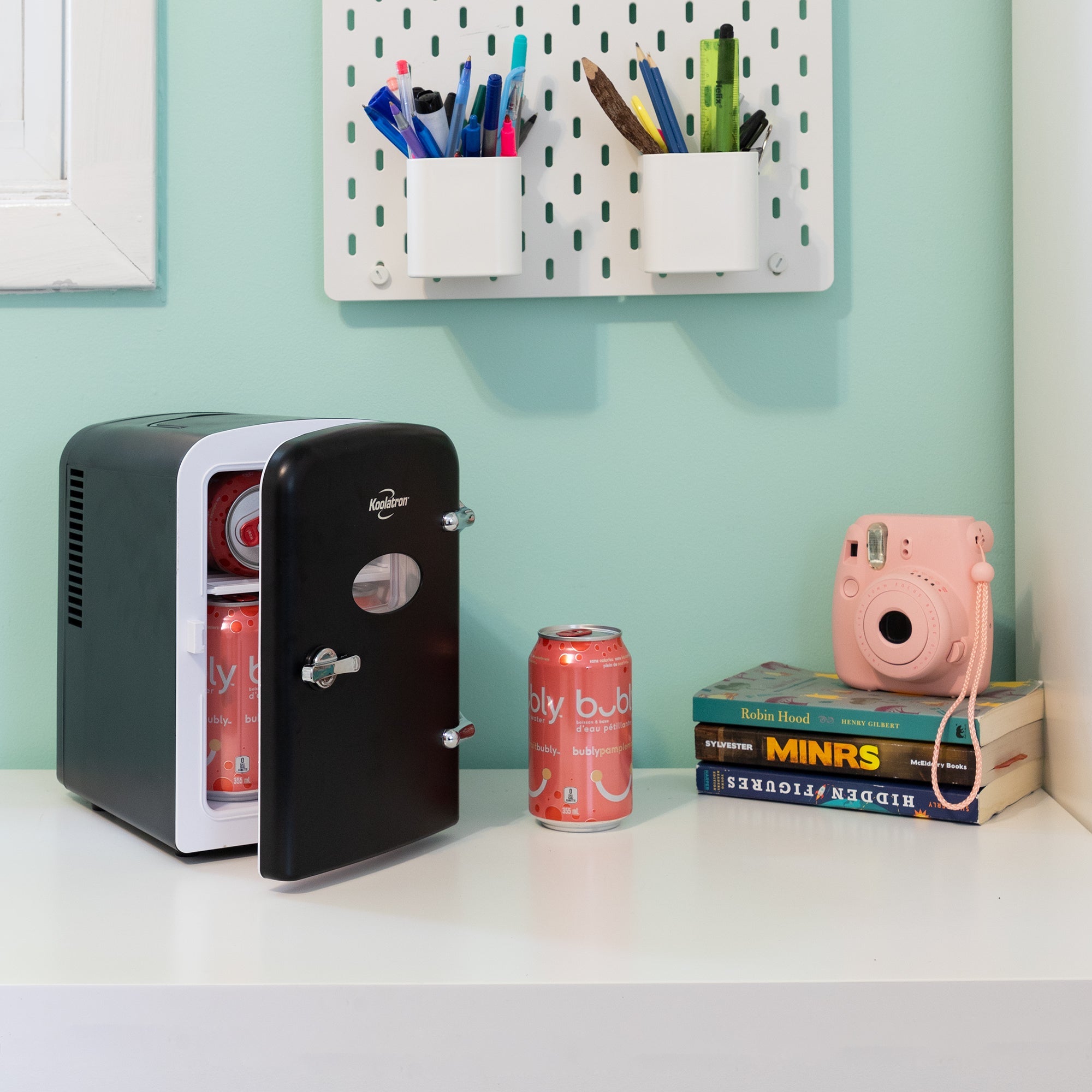 Lifestyle image of Kool-atronretro 6 can mini fridge, partly open with cans inside, on a white desktop with an aqua wall behind. There is a can of soda, a stack of books, and a pink camera to the right of the fridge and two cups of pens and pencils attached to the wall above