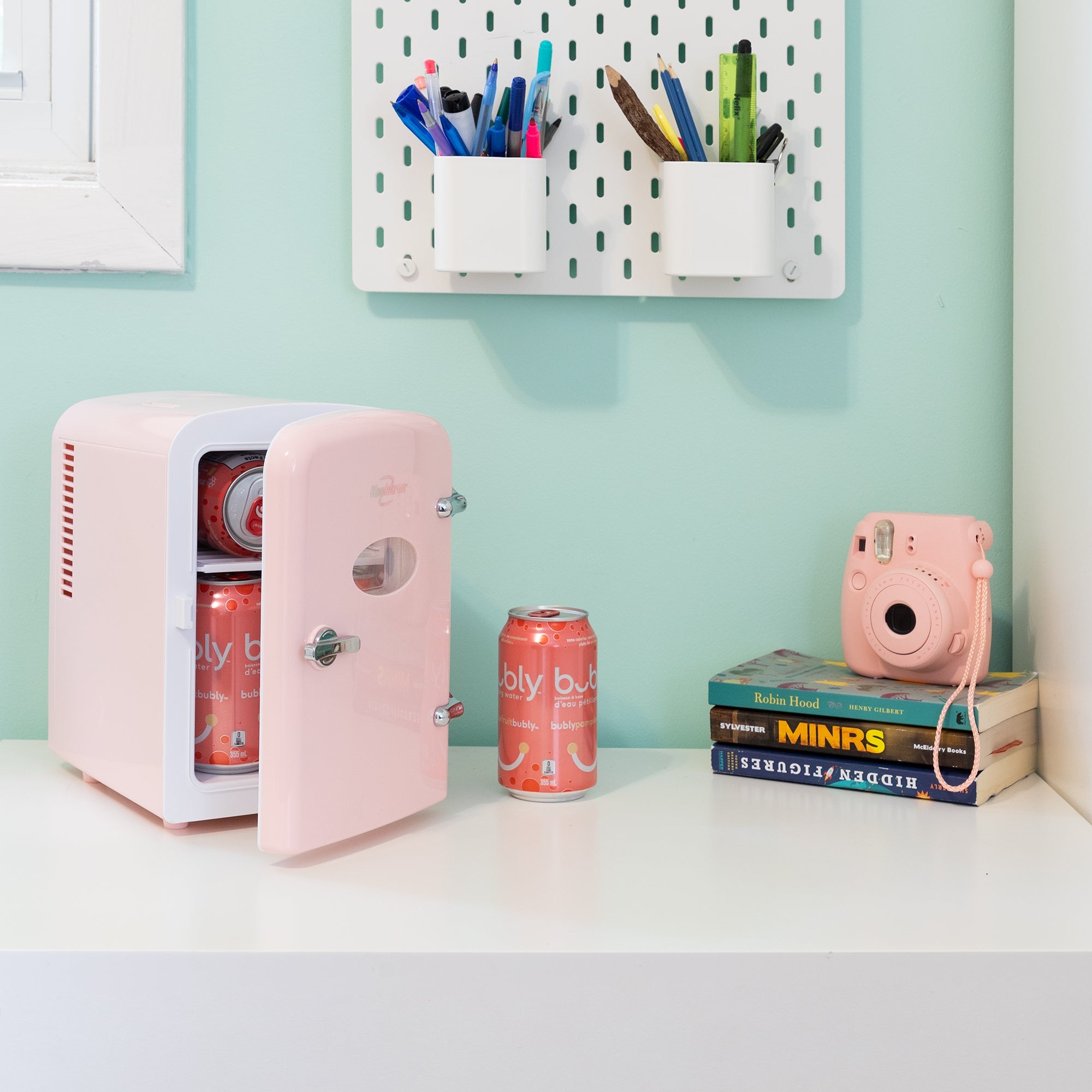 Lifestyle image of Kool-atronretro 6 can mini fridge, partly open with cans inside, on a white desktop with an aqua wall behind. There is a can of soda, a stack of books, and a pink camera to the right of the fridge and two cups of pens and pencils attached to the wall above