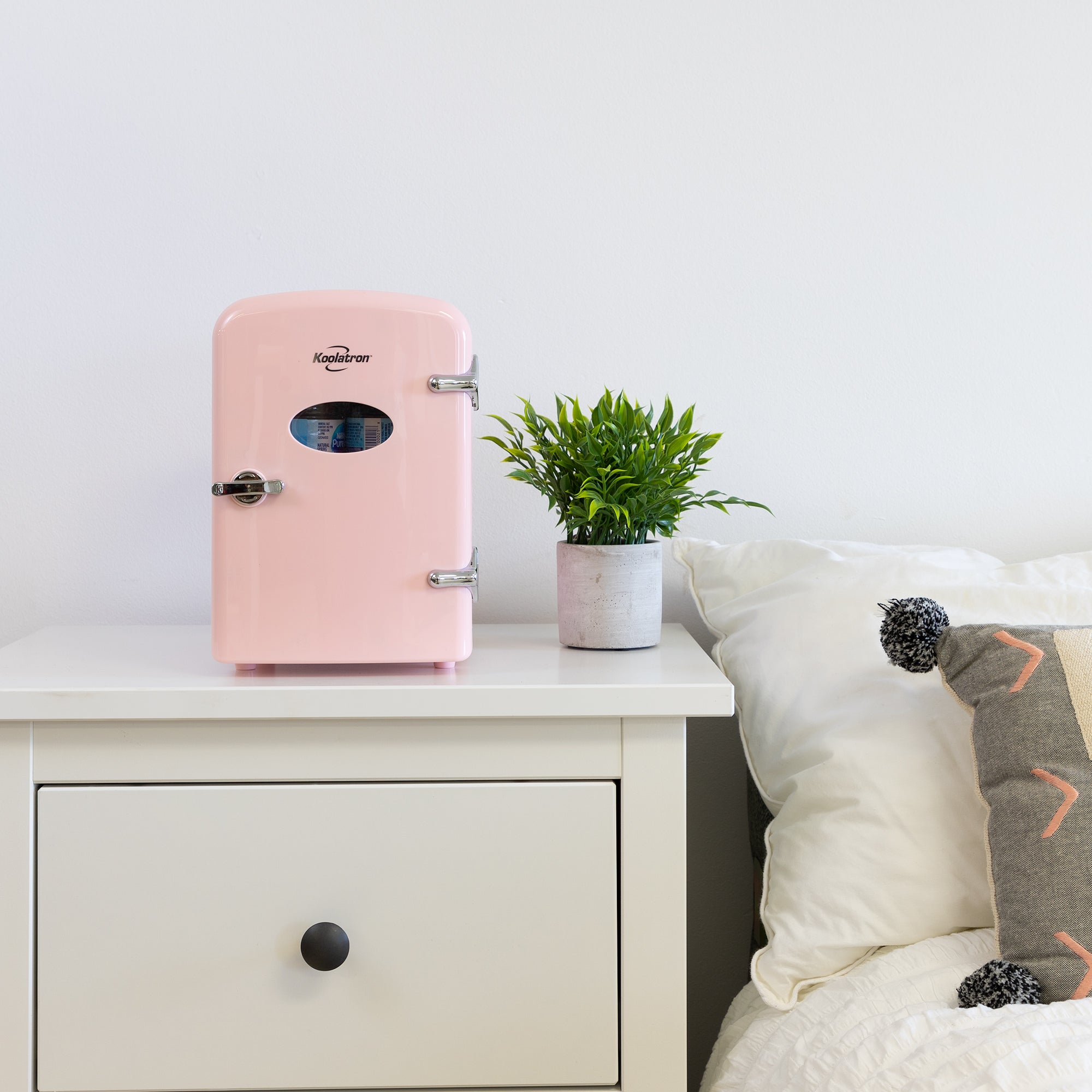Lifestyle image of Kool-atronretro 6 can mini fridge, closed, with a plant in a white pot on its right, on a white bedside table beside a bed with white linens